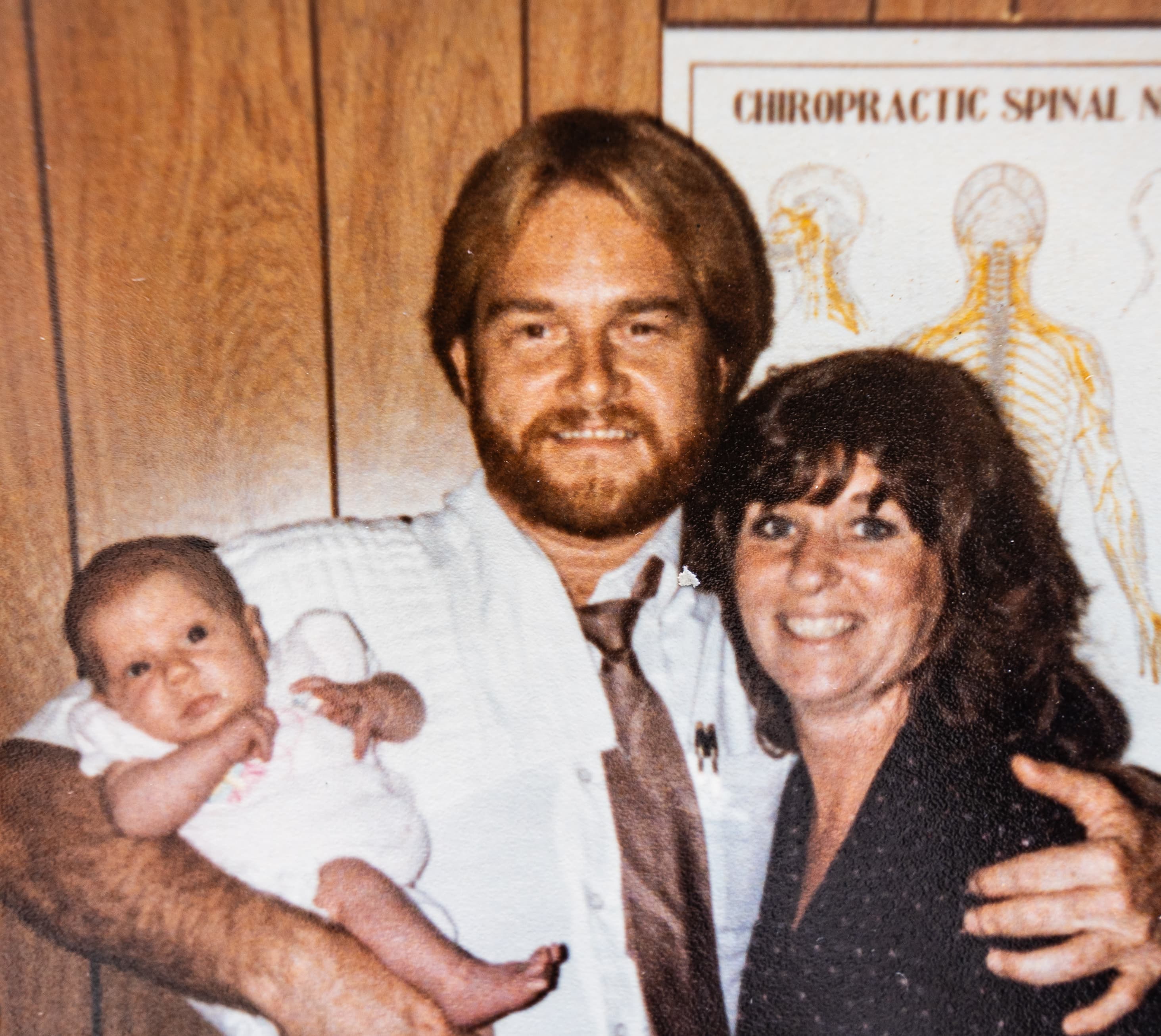 Three generations: baby Dr. Bridget held by her parents Dr. Bruce and Dr. Karen Edkin in front of a vintage chiropractic spinal poster